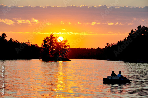 sunset on a Muskoka, Ontario lake with two women in silhouette in their paddleboat, © Joanne Dale