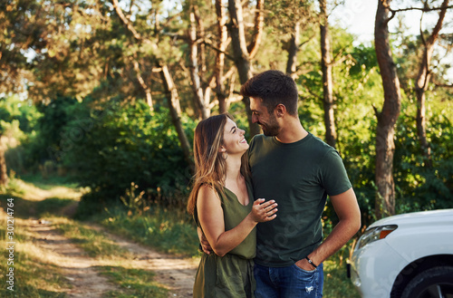 Standing near the white automobile. Beautiful young couple have a good time in the forest at daytime
