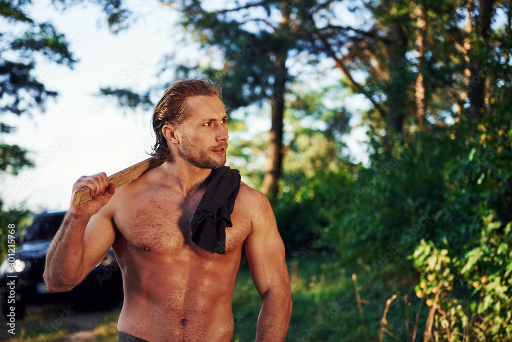 Close up portrait of woodsman with axe in hand. Handsome shirtless man with muscular body type is in the forest at daytime