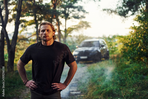 Black automobile behind. Portrait of man that stands in the forest and looks at nature