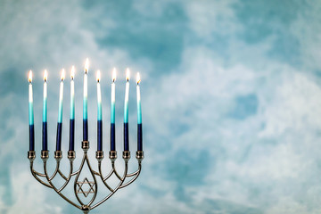 A silver menorah for the Jewish holiday Hanukkah with burning glowing eight candles on fire lit up on a blue background with copy space