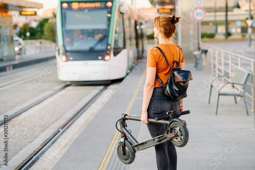 Back view of woman with backpack and folded electric scooter waiting at tram stop photo