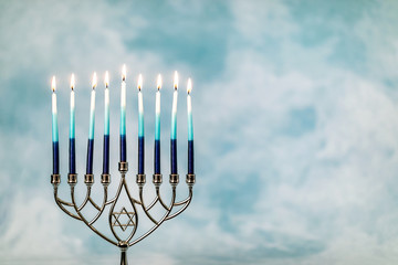 A silver menorah for the Jewish holiday Hanukkah with burning glowing eight candles on fire lit up on a blue background with copy space