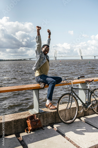 Young man with bicycle, sitting on railing by the sea, stretching photo