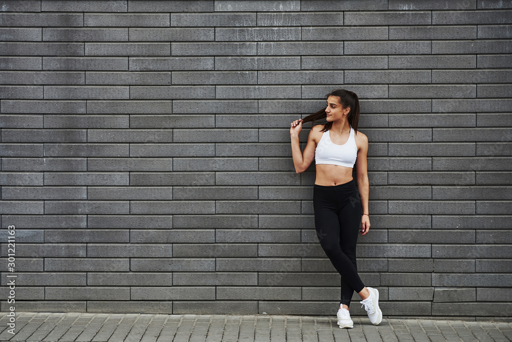 Young sportive brunette with slim body shape against brick wall in the city at daytime