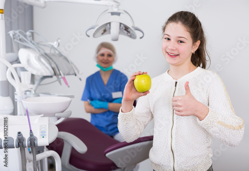 Girl with apple showing thumb up in dental clinic