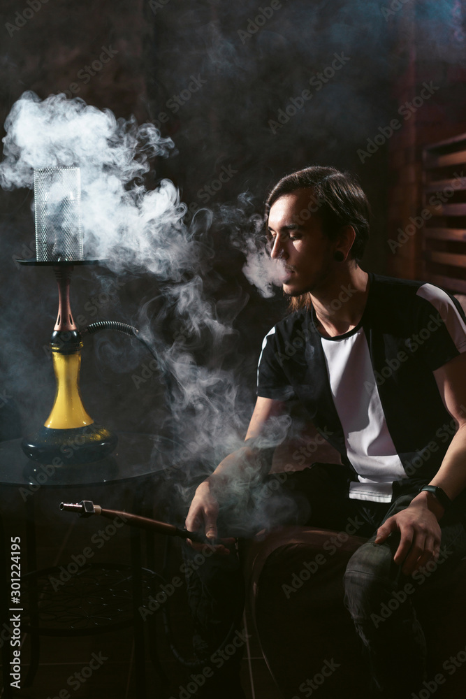 Portrait of man smoking traditional hookah pipe and making smoke clouds with shisha. Man exhaling smoke in hookah cafe or lounge Portrait of man smoking traditional hookah pipe and making smoke clouds with shisha. Man exhaling smoke in hookah cafe or lounge