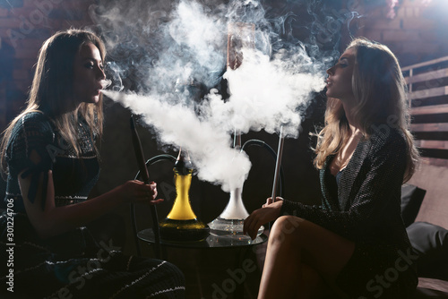 Girls party in hookah lounge. Group of two young women in backlight smoking shisha in cafe or bar, making smoke clouds, having fun, smiling. Relax concept. Friendship