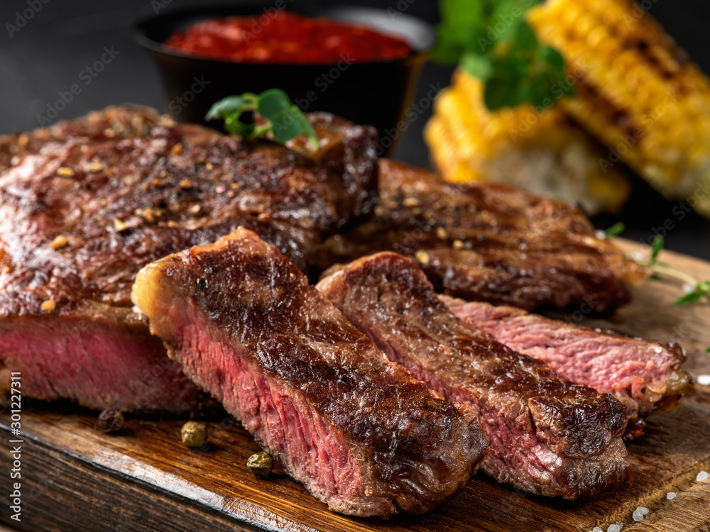 Sliced, fried, spiced steaks with herbs on wooden board, grilled corn, red sauce in small dark bowl on a black background. Close-up shot. Side view.