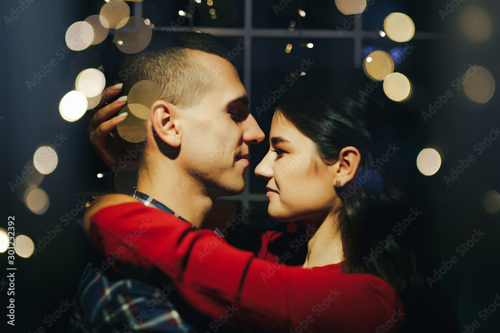 couple in love in the dark between the lights of garlands hugs and kisses. holiday and date for christmas. twelve hours, kiss on new year's eve. in the lights on the background of the window. couple in love in the dark between the lights of garlands hugs and kisses. holiday and date for christmas. twelve hours, kiss on new year's eve. in the lights on the background of the window.