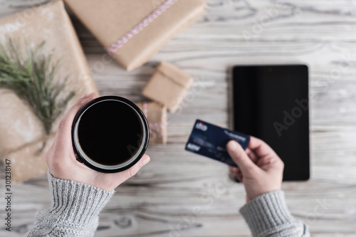 Fototapeta Naklejka Na Ścianę i Meble -  Woman with cup of coffee, credit card and tablet. Packing gifts. Cardboard box in craft paper on the rustic wood planks background. Christmas and New Year concept.