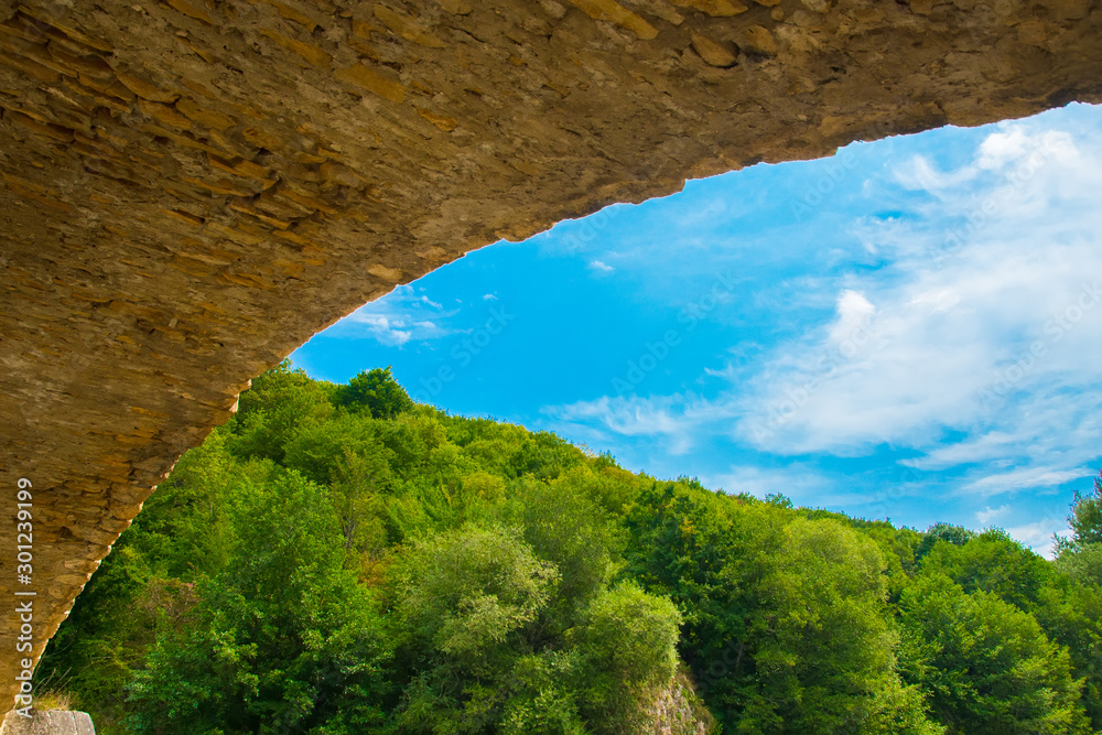 View of the blue sky and forest from under the arch of the stone bridge