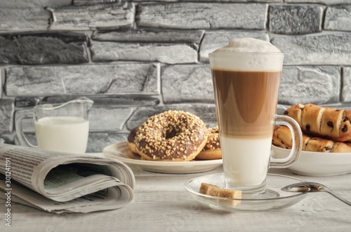 Conceptual delicious sweet breakfast. Latte macchiato coffee with sweet donuts with a hole and morning newspaper.