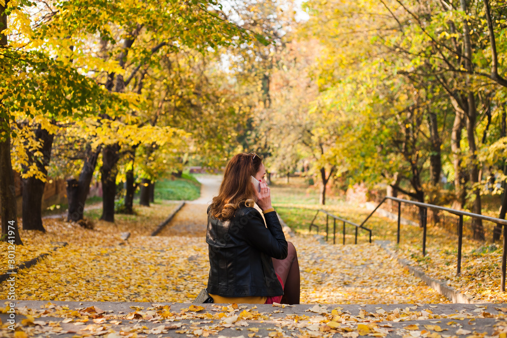 The beautiful young girl sitting on the stairs in the autumn park. Brunette in a leather jacket with a phone in her hands. The beautiful young girl sitting on the stairs in the autumn park. Brunette in a leather jacket with a phone in her hands.