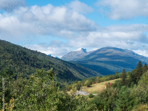 view on Romsdalen valley from the road E136 with snow capped peaks of mountains, green forest and small red yellow house. Blue sky white clouds background