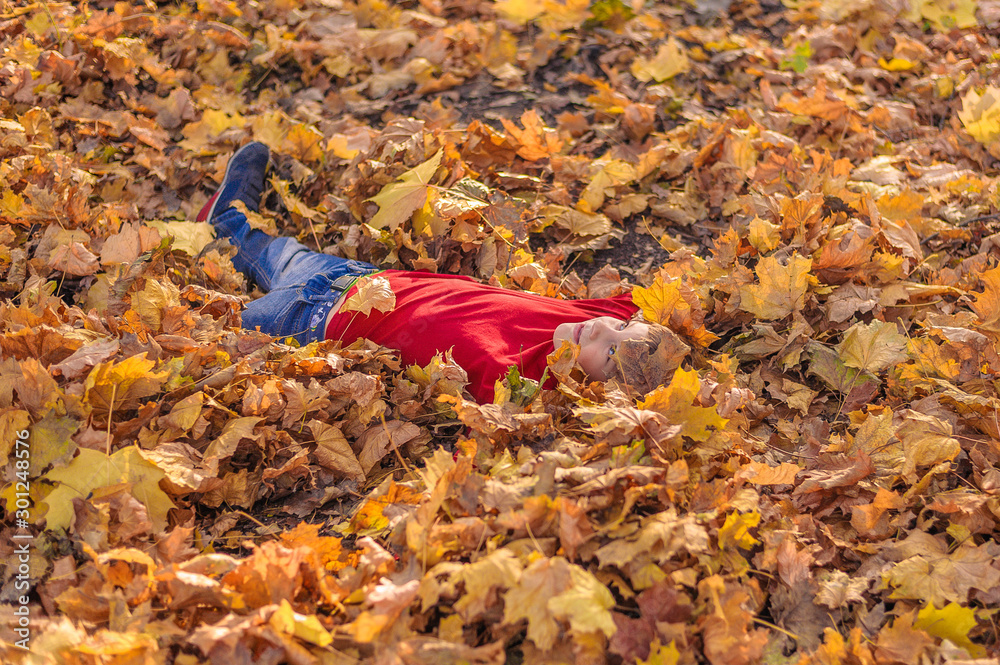 Obraz premium cheerful boy in a red jacket and blue jeans looks up to the sky, lying in yellow leaves in the autumn forest