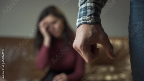 Angry Caucasian man at the foreground compressing his hand into fist. Young woman crying at the background and holding her head with hand. Domestic violence, tyranny, social problems. photo