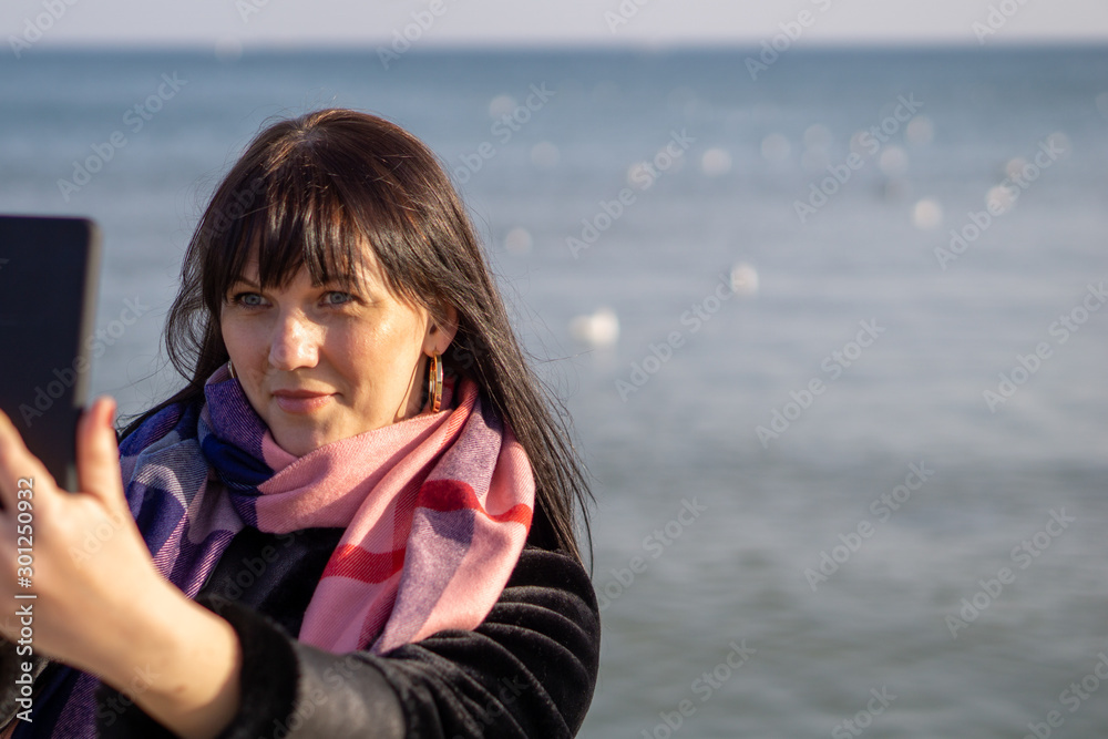 A young brunette woman in a black coat and pink scarf takes a picture of herself on the background of the sea on a sunny autumn day.