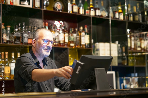 Professional elderly bartender in eyeglases registration of a new order by a cash register. Service concept.  photo