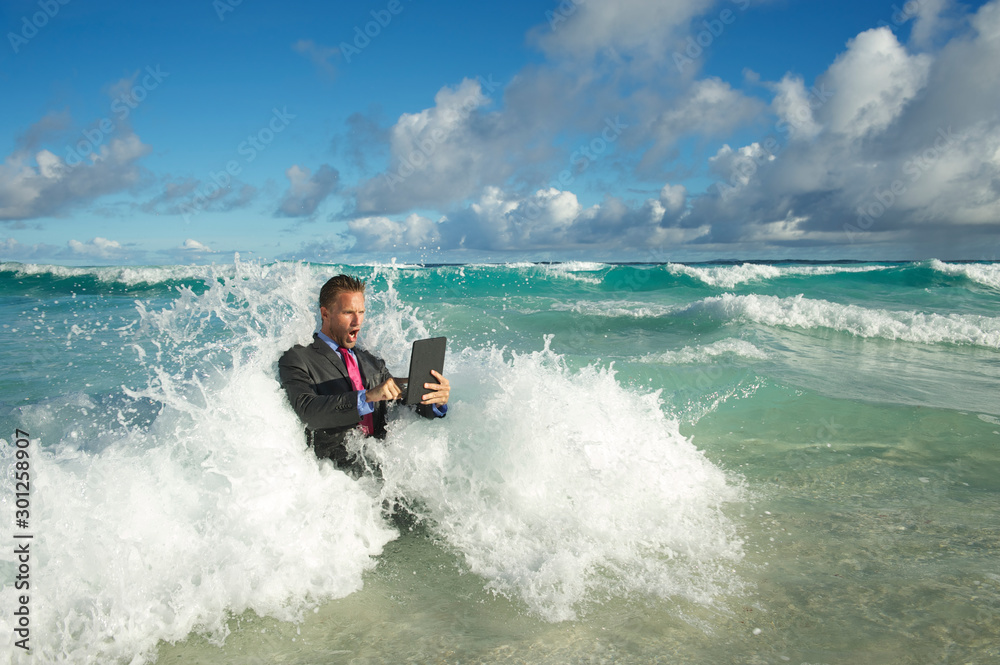Overwhelmed young businessman in a full suit having a rough time using his tablet computer to surf the web in crashing waves on the shore of a tropical beach Overwhelmed young businessman in a full suit having a rough time using his tablet computer to surf the web in crashing waves on the shore of a tropical beach
