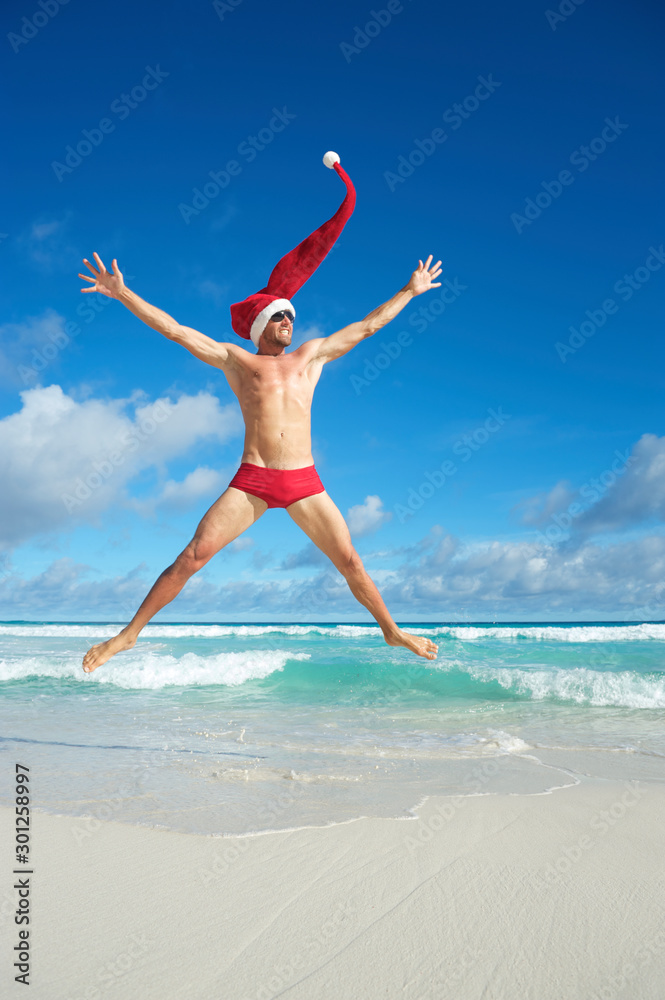 Thin Santa with extra long hat jumping in red swimming briefs on the shore of a beach in a tropical Christmas celebration Thin Santa with extra long hat jumping in red swimming briefs on the shore of a beach in a tropical Christmas celebration