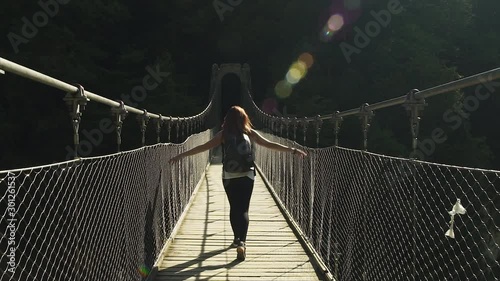 Female Explorer redhead girl walking, hiking, crossing beautiful bridge of Holzarte Footbridge, wooden metal grey bridge, black backpack. 4K UHD. Hands moving along grids, beautiful sun flare.