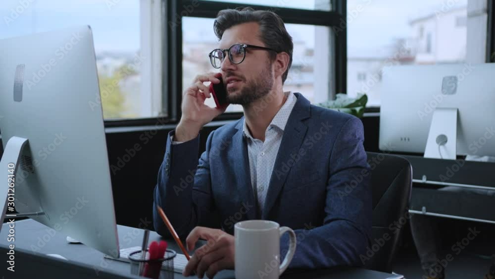 Office manager talking on smartphone with business partner or customer consulting phone call conversation sitting by computer in company workspace.