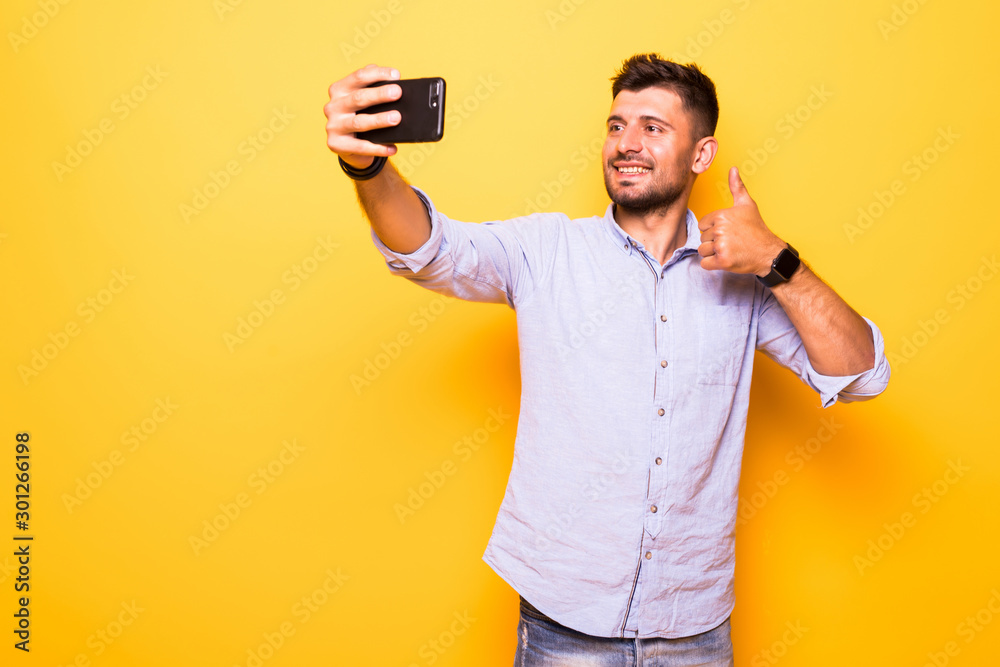 Portrait of a happy bearded man showing thumbs up gesture while taking a selfie over yellow background