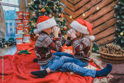 Full-body shot of beautiful children in santa hats and woolen sweaters, sitting near christmas tree, with a cup of cocoa, looking delighted and pleased. photo