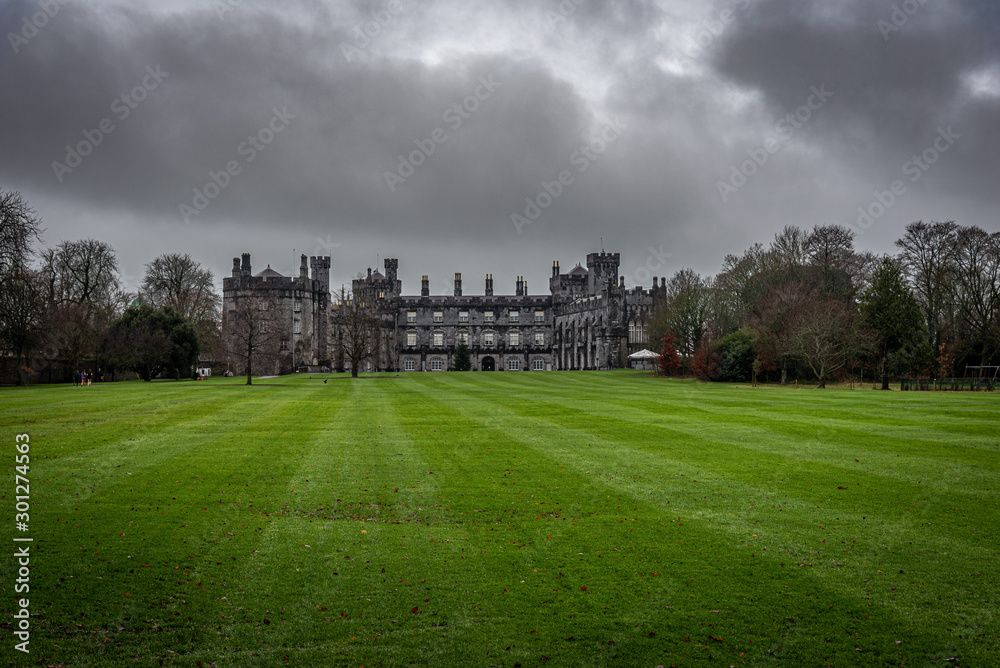 KILKENNY, IRELAND, DECEMBER 23, 2018: Kilkenny Castle seen from the huge garden on a dramatic cloudy day with dry leaves over the green grass while people run beside it.