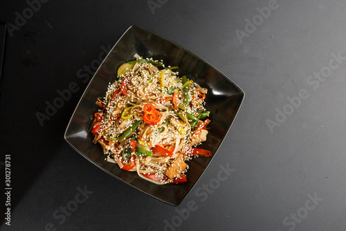 Chicken noodles bowl with vegetable. Udon stir-fry noodles with chicken meat and sesame in bowl on dark stone background copy space. Spaghetti pasta with chicken and vegetable. Horizontal photo.