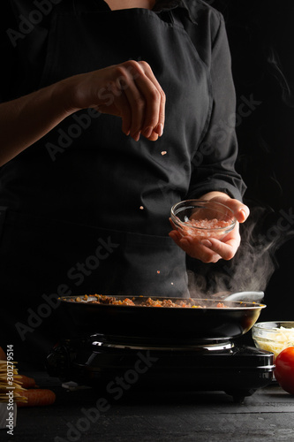 Chef fries vegetables with meat for cooking a delicious dish. Salt with salt. On a black background. Vertical frame.