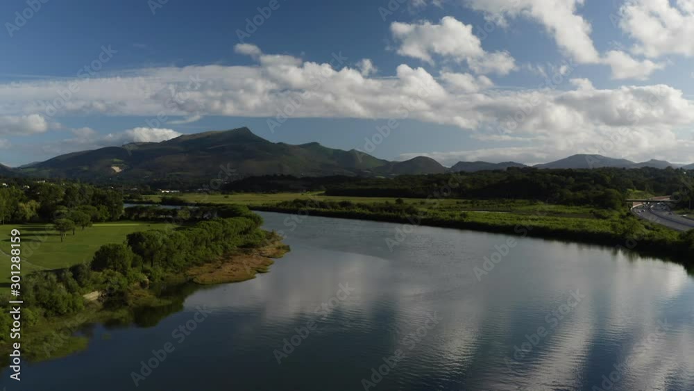 Aerial Drone View of beautiful river with water reflections near french mountains in Basque country, blue sky, clouds and trees landscape, valley, volcanic, green nature, 4K UHD. Tracking out. Aerial Drone View of beautiful river with water reflections near french mountains in Basque country, blue sky, clouds and trees landscape, valley, volcanic, green nature, 4K UHD. Tracking out.