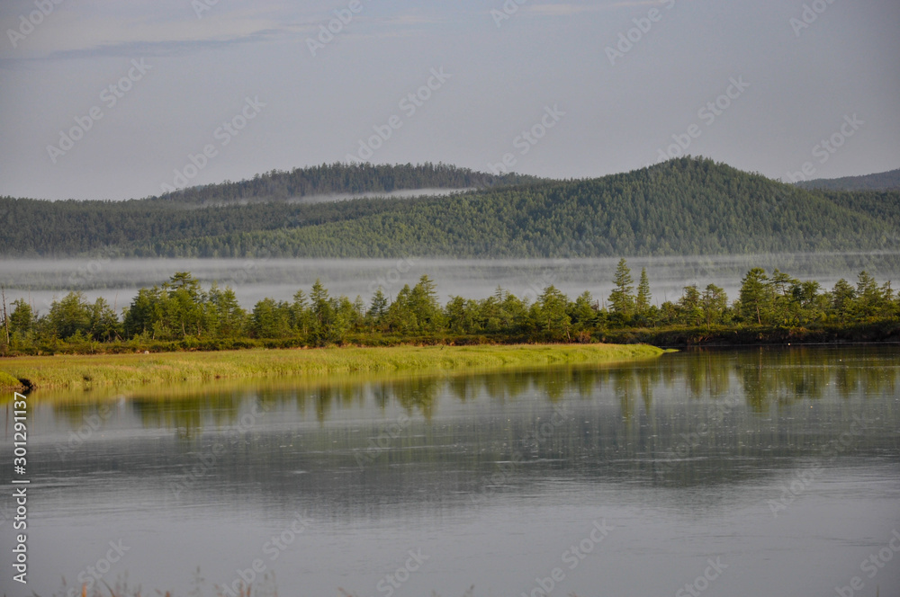 cloudy day, fog, forest and mountains are reflected in the gray water of the river.