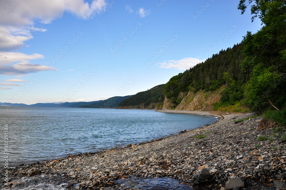 The coast of the sea. Stone spit, mountains covered by a forest, rocks, blue sky.
