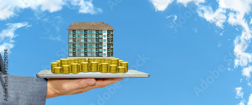  Businessman holding a tablet with piles of golden coins and condominium on white cloud and blue sky background