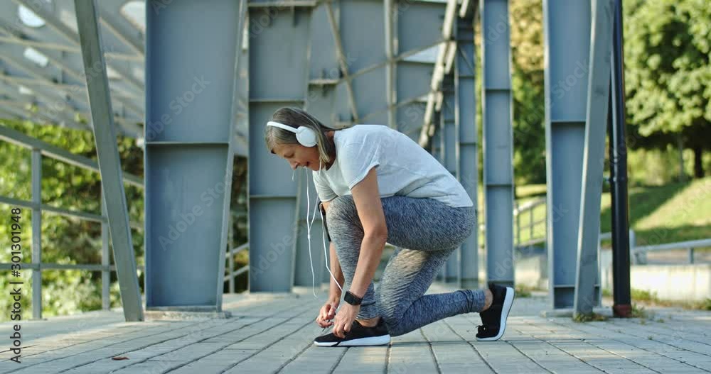 Caucasian senior sporty woman in headphones leaning down and tying the shoe laces while stops during jogging. Outdoor. Caucasian senior sporty woman in headphones leaning down and tying the shoe laces while stops during jogging. Outdoor.