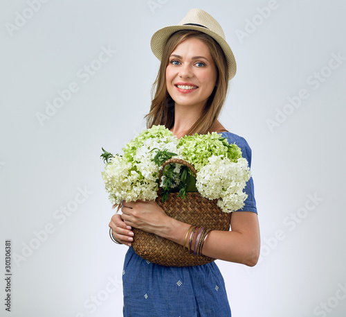 Smiling woman wearing hat holding straw bag with flowers.