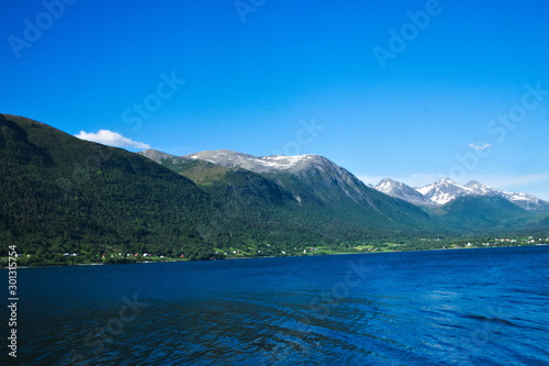 Isfjorden fjord and adjacent mountains with snow near Andalsnes, Norway, with homes near the shoreline and clear blue sky in the background on a sunny day.