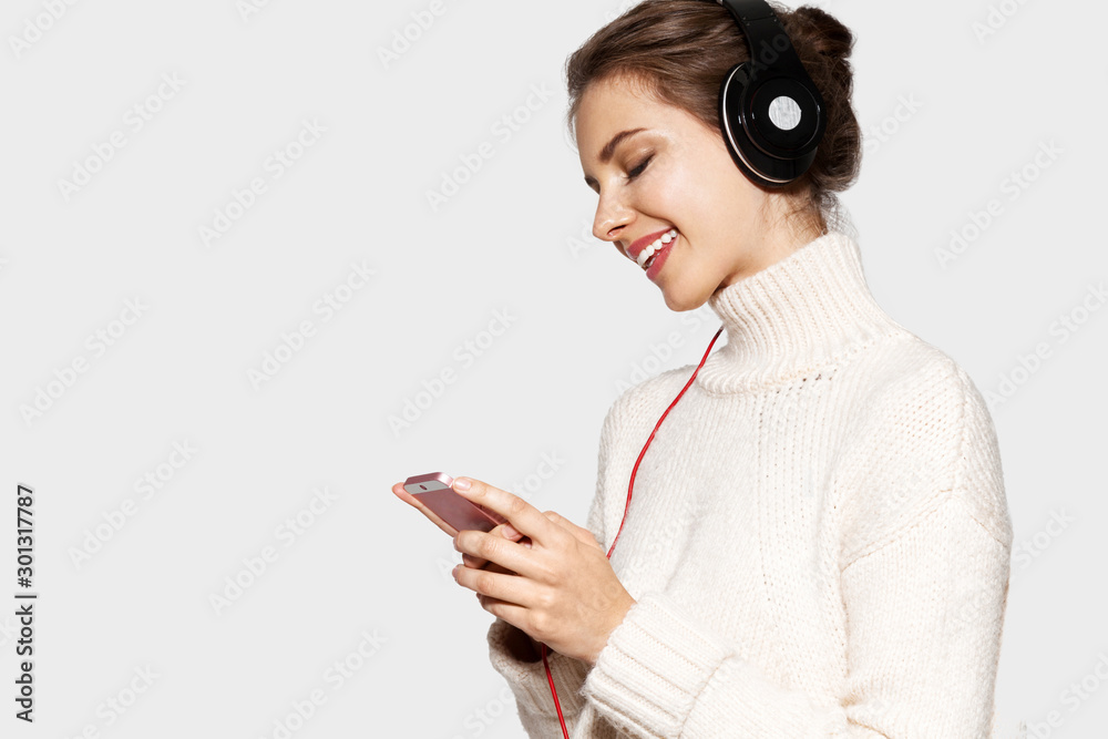 Portrait of happy young woman holding iphone and wearing headphones. Pretty lady listening to music and posing on grey background. Copy space in left side Portrait of happy young woman holding iphone and wearing headphones. Pretty lady listening to music and posing on grey background. Copy space in left side