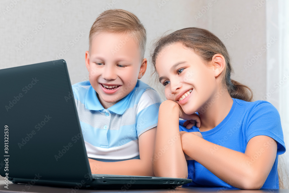 Children at the computer. Little cute boy and little beautiful girl look at the laptop screen and smile cute. Distance learning, fun leisure concept. Happy children use a laptop. Children at the computer. Little cute boy and little beautiful girl look at the laptop screen and smile cute. Distance learning, fun leisure concept. Happy children use a laptop.