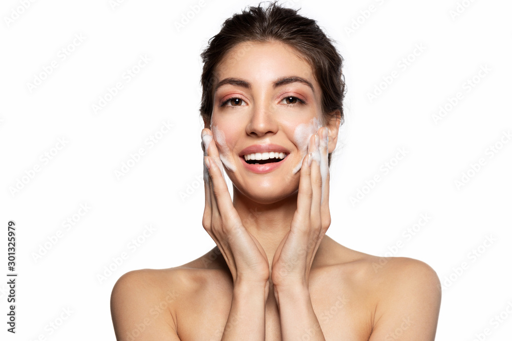Happy smiling young woman cleaning face with soap foam studio portrait. Pretty girl massaging skin on cheeks. Face skincare natural cosmetic. Morning routine, facial beauty treatment. White copy space Happy smiling young woman cleaning face with soap foam studio portrait. Pretty girl massaging skin on cheeks. Face skincare natural cosmetic. Morning routine, facial beauty treatment. White copy space