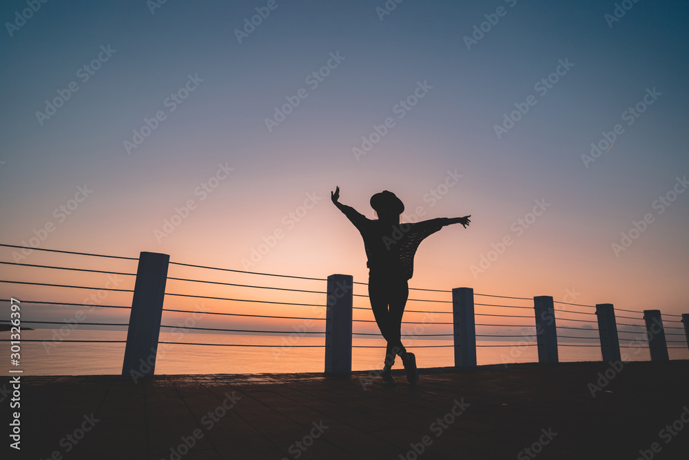 Fototapeta premium Silhouette of one lonely free carefree traveler woman in hat with open outstretched arms standing on sea pier at sunset. Happy freedom concept and enjoying life