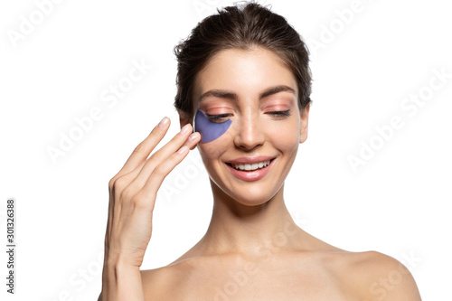 Happy smiling young woman touching patch under eye studio portrait. Beautiful girl apply moisturizing skin, smoothing out fine wrinkles, combating bruises and dark circles under eyes. White copy space