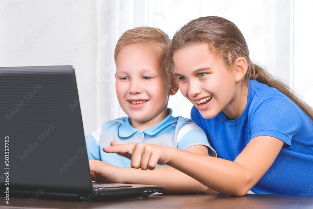 Two beautiful cute happy smiling children, a boy and a girl, use laptop for distance learning or entertainment. Children look at the screen with interest and the girl points with the finger to display Two beautiful cute happy smiling children, a boy and a girl, use laptop for distance learning or entertainment. Children look at the screen with interest and the girl points with the finger to display