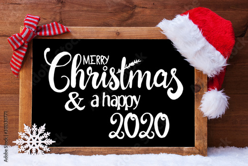 Chalkboard With English Calligraphy Merry Christmas And A Happy 2020. Christmas Decoration Like Santa Hat And Bow. Wooden Background With Snow