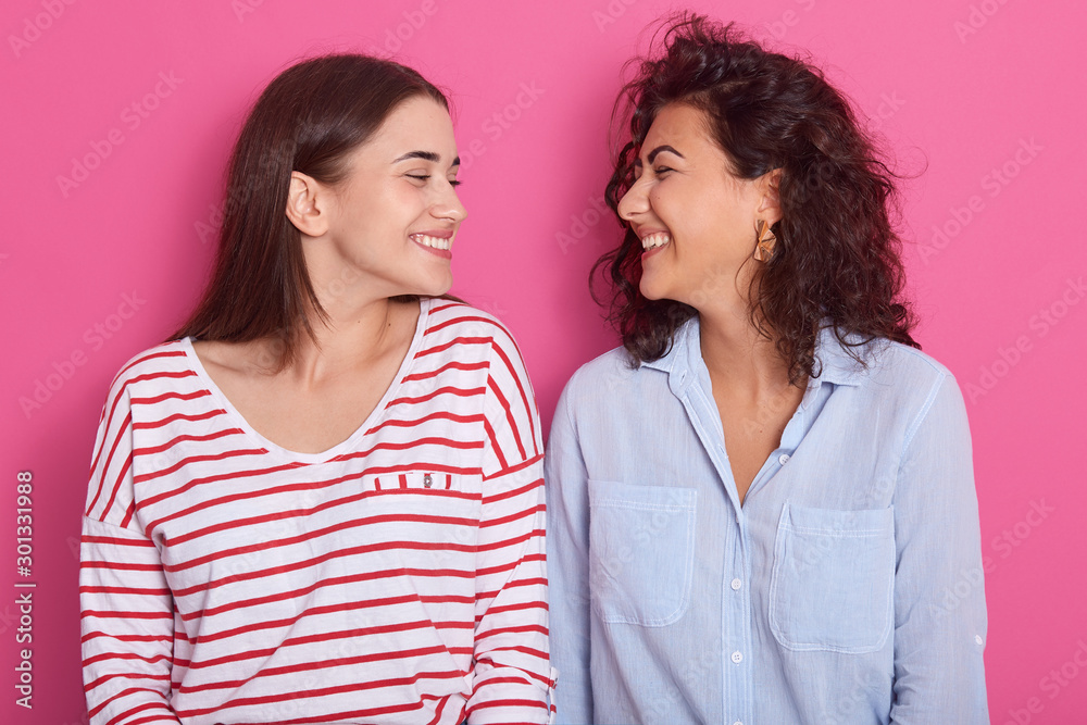 Close up portrait of two beautiful young women posing over pink background, friends wearing casually clothing, standing and look smiling at each other, dark haired females. Friendship concept. Close up portrait of two beautiful young women posing over pink background, friends wearing casually clothing, standing and look smiling at each other, dark haired females. Friendship concept.