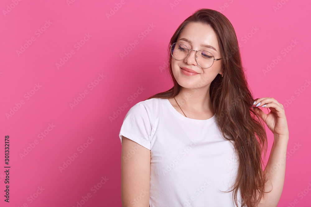 Portrait of happy young cute brown haired girl sytands smiling and looking aside, touches hair, posing isolated on pink background, looks thouthfull, dreams about something. Copyspace for advertisment Portrait of happy young cute brown haired girl sytands smiling and looking aside, touches hair, posing isolated on pink background, looks thouthfull, dreams about something. Copyspace for advertisment
