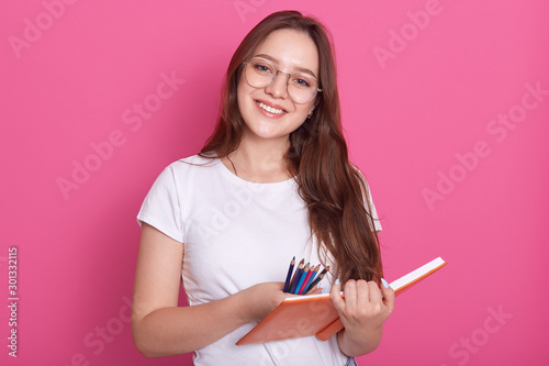 People, youth, lifestyle and education concept. Close up portrait of beautiful student girl ready to make notes in copybook, having pleasant look, holding organaizer and colored pencils in hands. photo