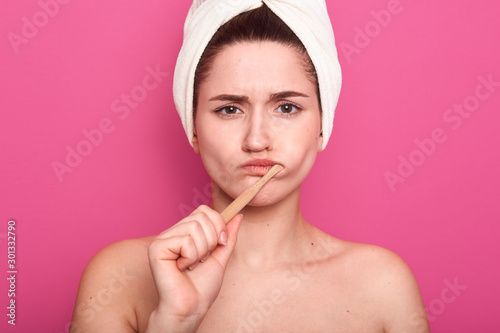 Close up portrait of attractive caucasian woman with dark hair posing isolated over pink background, femaklw with bare shouldersand white towel on head, lady looking at camera, brushing teeth. photo